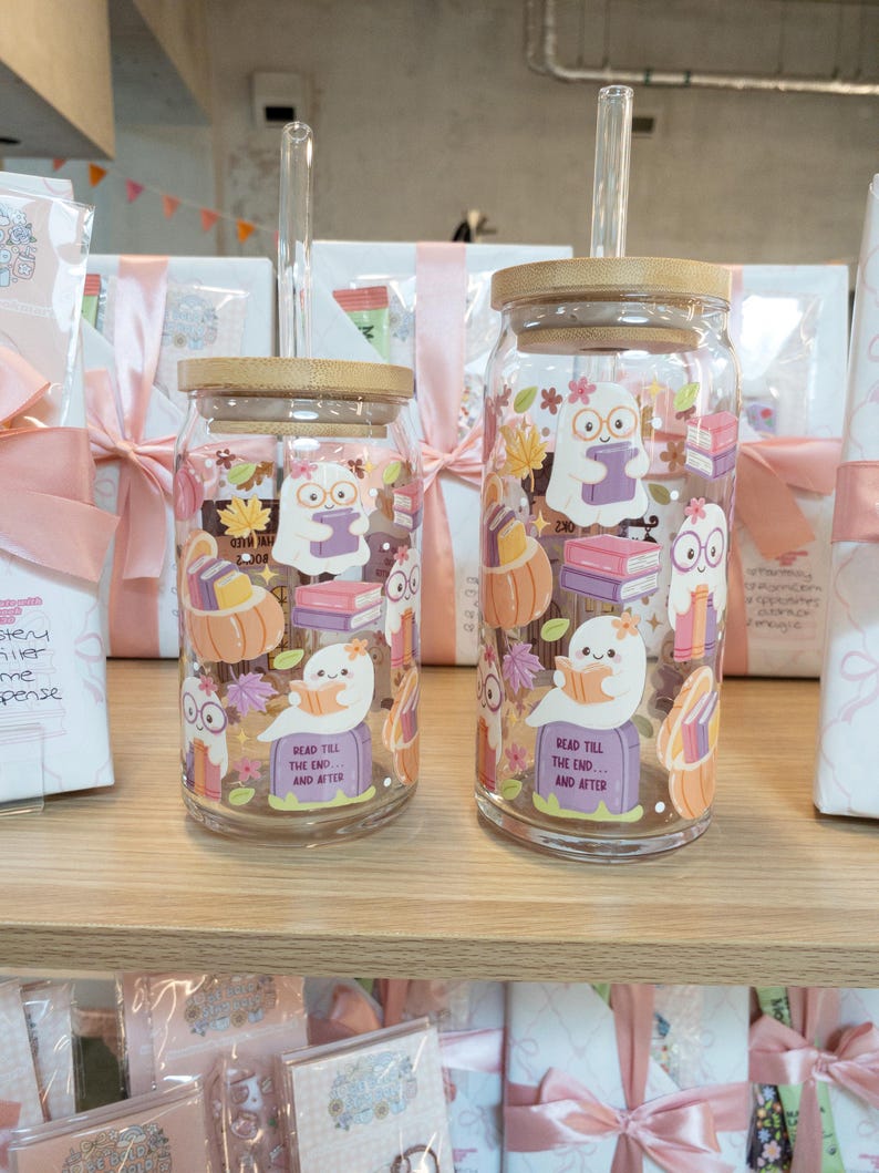 May include: Two clear glass tumblers with bamboo lids and glass straws. The tumblers feature a whimsical design of a ghost reading a book, pumpkins, and fall leaves. The text "Read Till The End... And After" is printed on the bottom.