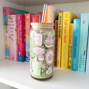 May include: A clear glass tumbler with a bamboo lid and a clear straw, decorated with pastel-colored illustrations of books, cakes, and flowers. The tumbler sits on a white shelf with a collection of books in various colors.