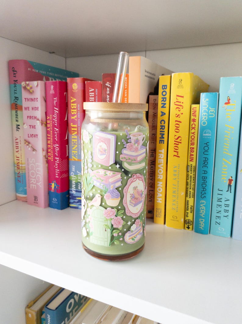 May include: A clear glass tumbler with a bamboo lid and a clear straw, decorated with pastel-colored illustrations of books, cakes, and flowers. The tumbler sits on a white shelf with a collection of books in various colors.