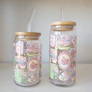 May include: Two clear glass tumblers with bamboo lids and glass straws. The tumblers are decorated with pastel-colored illustrations of books, cakes, flowers, and birds. The tumblers are sitting on a white surface.