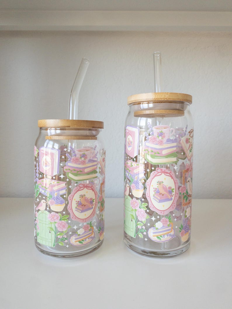 May include: Two clear glass tumblers with bamboo lids and glass straws. The tumblers are decorated with pastel-colored illustrations of books, cakes, flowers, and birds. The tumblers are sitting on a white surface.