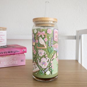 May include: A glass tumbler with a bamboo lid and a clear straw. The tumbler is decorated with a whimsical garden scene featuring birds, teacups, and flowers in shades of pink, green, and purple. The tumbler is sitting on a wooden surface.