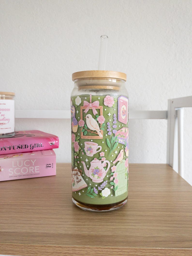 May include: A glass tumbler with a bamboo lid and a clear straw. The tumbler is decorated with a whimsical garden scene featuring birds, teacups, and flowers in shades of pink, green, and purple. The tumbler is sitting on a wooden surface.