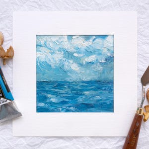 Puede incluir: Pintura al óleo original del océano y el cielo en tonos azules y blancos, presentada en un passe-partout blanco. Un tubo de pintura azul, una espátula y conchas marinas están dispuestas alrededor de la obra de arte.