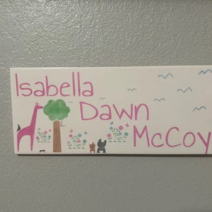 May include: A rectangular white sign with the names "Isabella Dawn McCoy" in pink script. The sign features a pink giraffe, a green tree, and small animal figures, with blue birds and flowers.