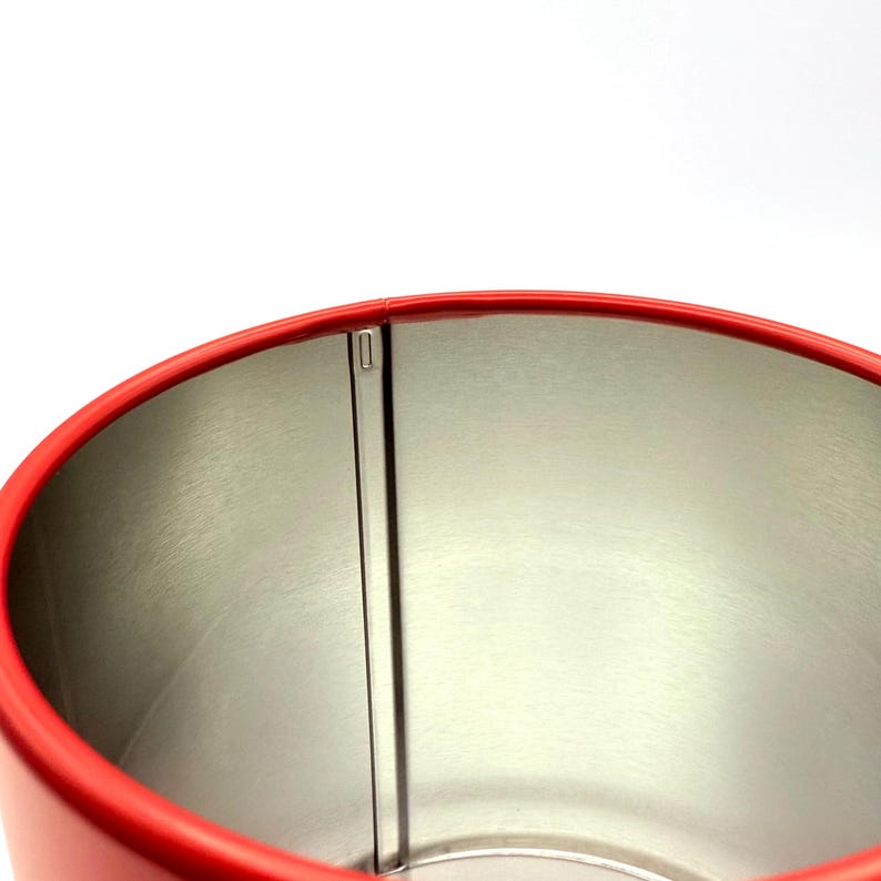 May include: A cylindrical metal container with a bright red exterior and a metallic silver interior. The top rim is finished in red, and a vertical seam runs down the inside wall, showing the construction of the canister's metal body.