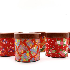 May include: A collection of six cylindrical canisters with dark brown wooden lids. Each canister features a vibrant red base decorated with various colourful floral patterns, including small blossoms and intricate petal designs in shades of pink, white, yellow, and blue.