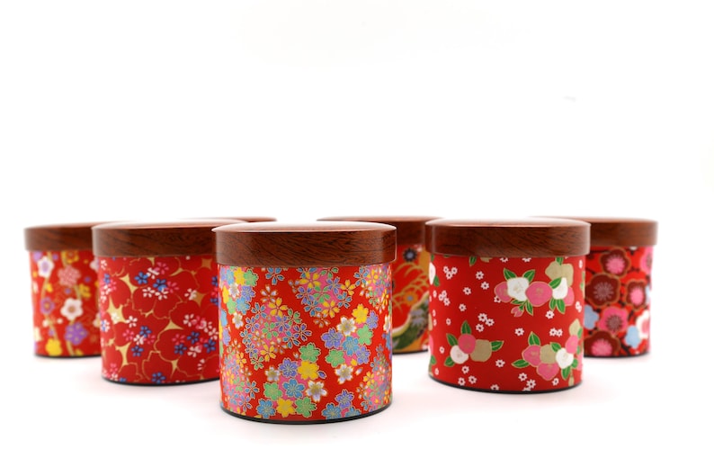 May include: A collection of six cylindrical canisters with dark brown wooden lids. Each canister features a vibrant red base decorated with various colourful floral patterns, including small blossoms and intricate petal designs in shades of pink, white, yellow, and blue.