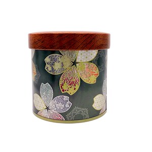 May include: A cylindrical metal storage canister featuring a dark green background decorated with a repeating pattern of stylised cherry blossom petals in shades of pink, white, and yellow. The container has a round, wood-grain patterned lid.