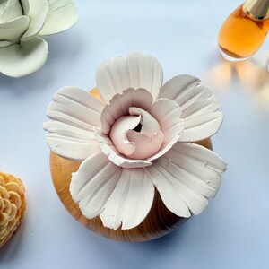May include: A white ceramic flower diffuser with a pale pink centre, mounted on a round, light brown wood-grain base. The flower features five large, textured petals surrounding a layered, ruffled core, creating a decorative scent diffuser.