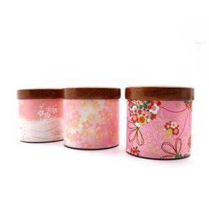 May include: Three cylindrical containers with dark brown wooden lids. Each container features a distinct pink patterned exterior, including floral motifs, cherry blossom designs, and intricate decorative line work on a light pink background.