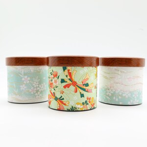 May include: Three cylindrical storage canisters with dark brown wooden lids. The two outer canisters feature a light mint green background with white cherry blossom patterns. The centre canister has a light green base with red and gold ribbons.