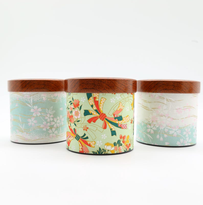 May include: Three cylindrical storage canisters with dark brown wooden lids. The two outer canisters feature a light mint green background with white cherry blossom patterns. The centre canister has a light green base with red and gold ribbons.
