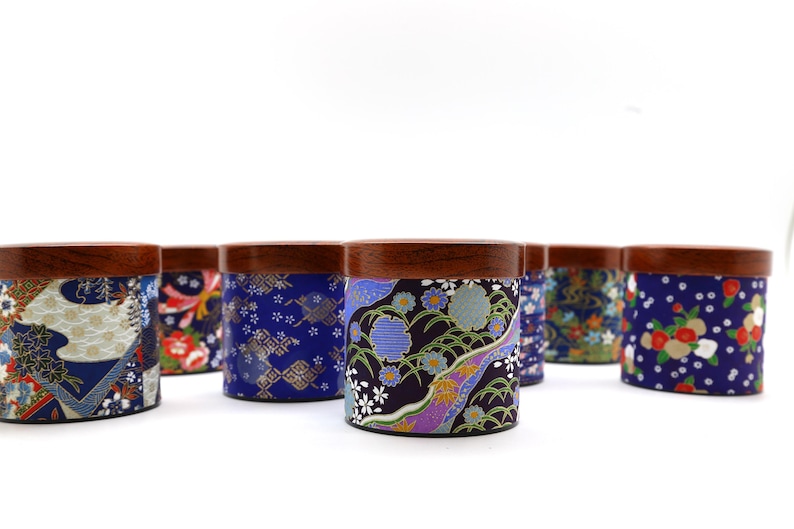 May include: A collection of cylindrical containers with dark brown wooden lids, each wrapped in fabric featuring diverse blue, purple, and red floral and geometric patterns. The containers are arranged in a group against a white background.