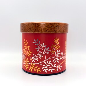 May include: A cylindrical red storage canister featuring a wooden lid with a visible grain pattern. The exterior of the canister is decorated with a botanical print of leaves and berries in shades of yellow, white, and metallic silver.