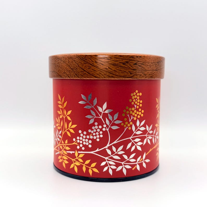 May include: A cylindrical red storage canister featuring a wooden lid with a visible grain pattern. The exterior of the canister is decorated with a botanical print of leaves and berries in shades of yellow, white, and metallic silver.