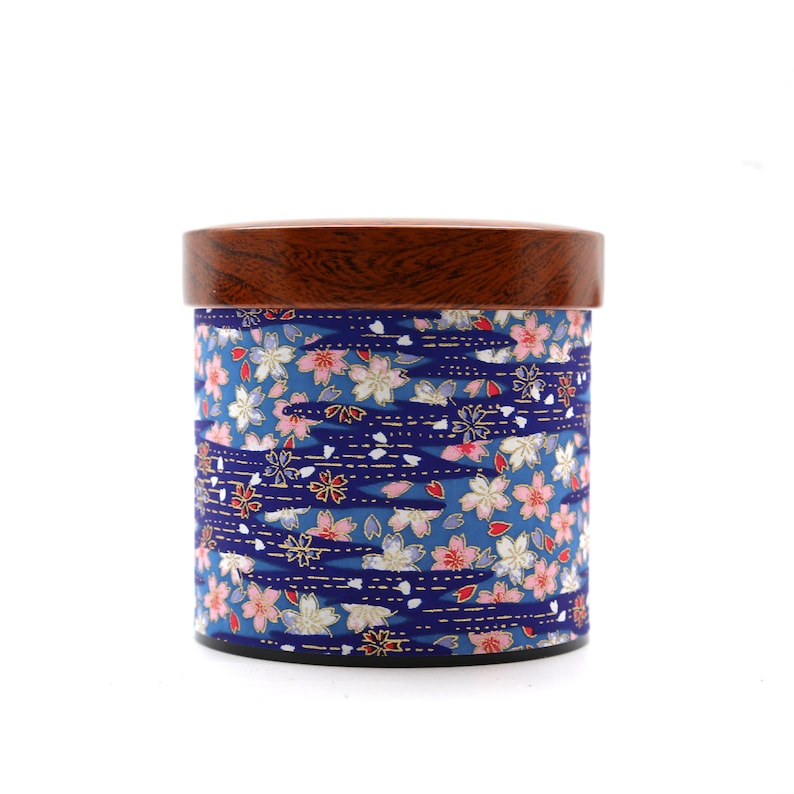 May include: A cylindrical storage canister featuring a dark brown wooden lid and a blue body decorated with a repeating pattern of pink and white cherry blossoms interspersed with dark blue wavy lines and small gold accents throughout the design.