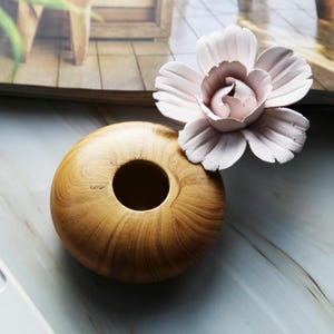 May include: A wooden bowl with a round opening in the centre. The bowl is light brown with a wood grain pattern. A pink flower is sitting on top of the bowl.