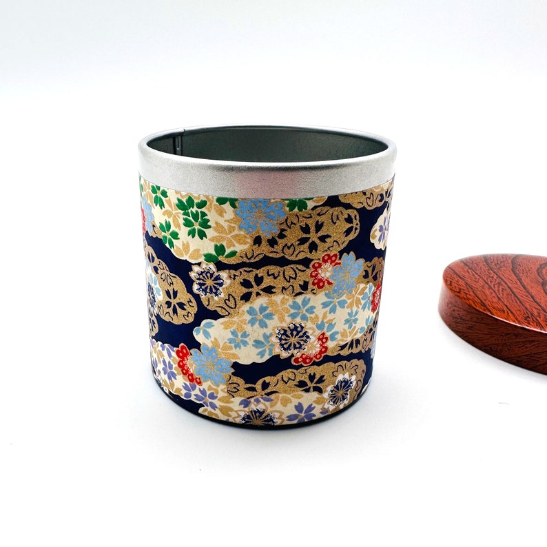 May include: A cylindrical metal tea canister featuring a dark blue background with a repeating pattern of gold, light blue, red, and green floral motifs. The canister has a silver metal rim and is accompanied by a separate, round, dark wood-grain lid.