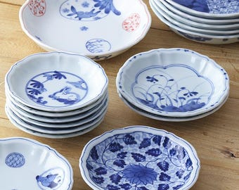 May include: A collection of small, round ceramic plates featuring scalloped edges and various blue and white patterns, including floral motifs, rabbits, and geometric designs. Some plates display additional red accents on the white surface.