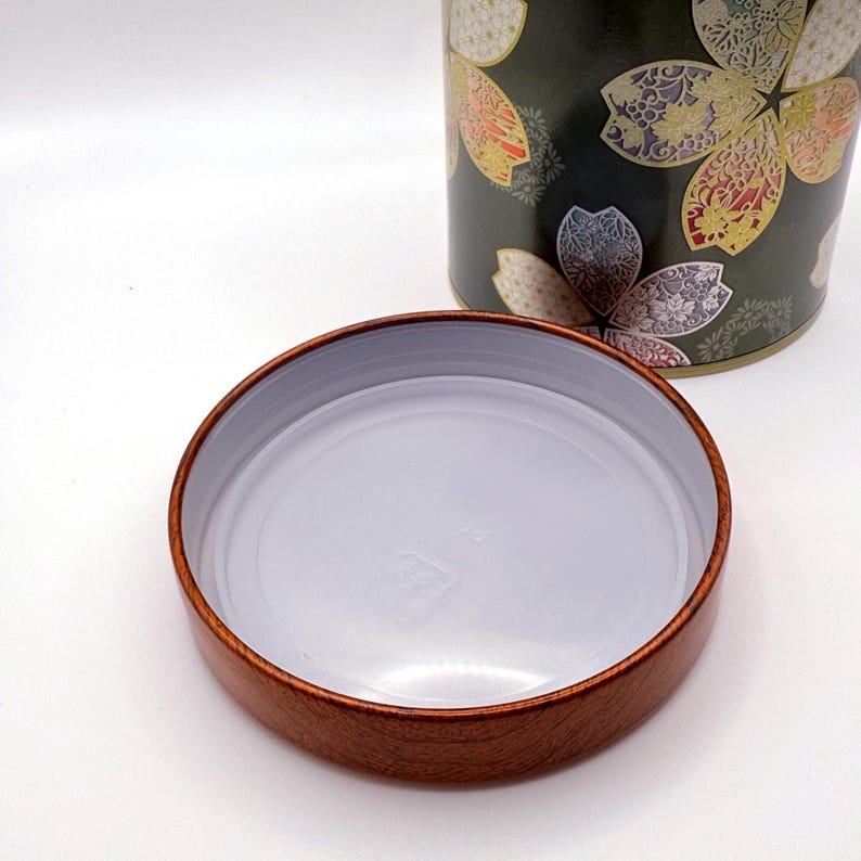 May include: A cylindrical metal tea canister featuring a dark green background with a gold, white, and pink floral pattern. The canister is accompanied by a separate, round lid that has a wood-grain exterior finish and a smooth white interior.