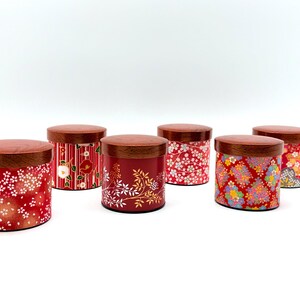 May include: A collection of six cylindrical storage canisters with dark brown wooden lids. Each canister features a distinct red-based exterior with varied floral and geometric patterns, including cherry blossoms, camellias, and intricate gold-toned motifs.