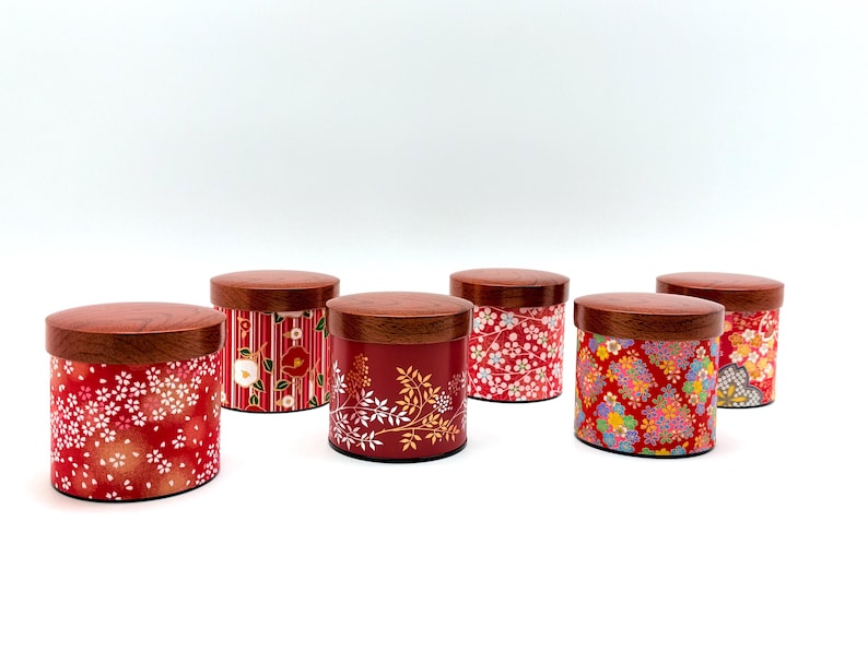 May include: A collection of six cylindrical storage canisters with dark brown wooden lids. Each canister features a distinct red-based exterior with varied floral and geometric patterns, including cherry blossoms, camellias, and intricate gold-toned motifs.