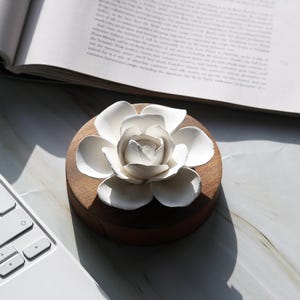 May include: A white ceramic flower diffuser sits on a wooden base. The flower has five petals and is designed to hold essential oils.