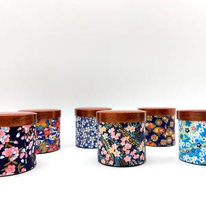 May include: A collection of six cylindrical storage canisters with dark wood lids. Each canister features a unique fabric wrap with colourful floral patterns, including cherry blossoms and geometric designs in shades of blue, pink, and gold.