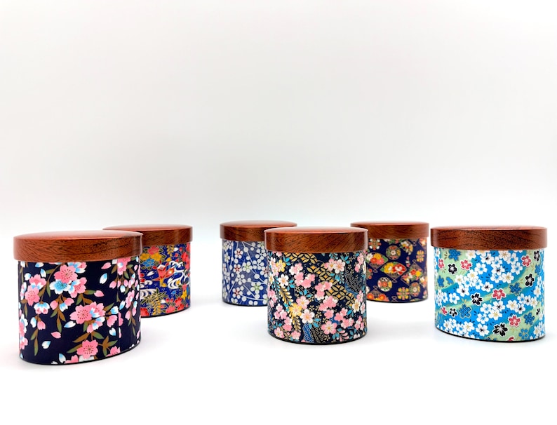 May include: A collection of six cylindrical storage canisters with dark wood lids. Each canister features a unique fabric wrap with colourful floral patterns, including cherry blossoms and geometric designs in shades of blue, pink, and gold.