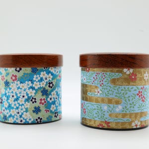 May include: Two cylindrical canisters with dark brown wooden lids. The left canister features a dense pattern of blue, white, and pink cherry blossoms on a light green background. The right canister displays a light blue floral pattern with gold accents.