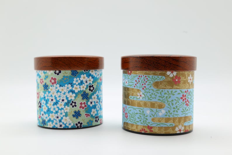 May include: Two cylindrical canisters with dark brown wooden lids. The left canister features a dense pattern of blue, white, and pink cherry blossoms on a light green background. The right canister displays a light blue floral pattern with gold accents.