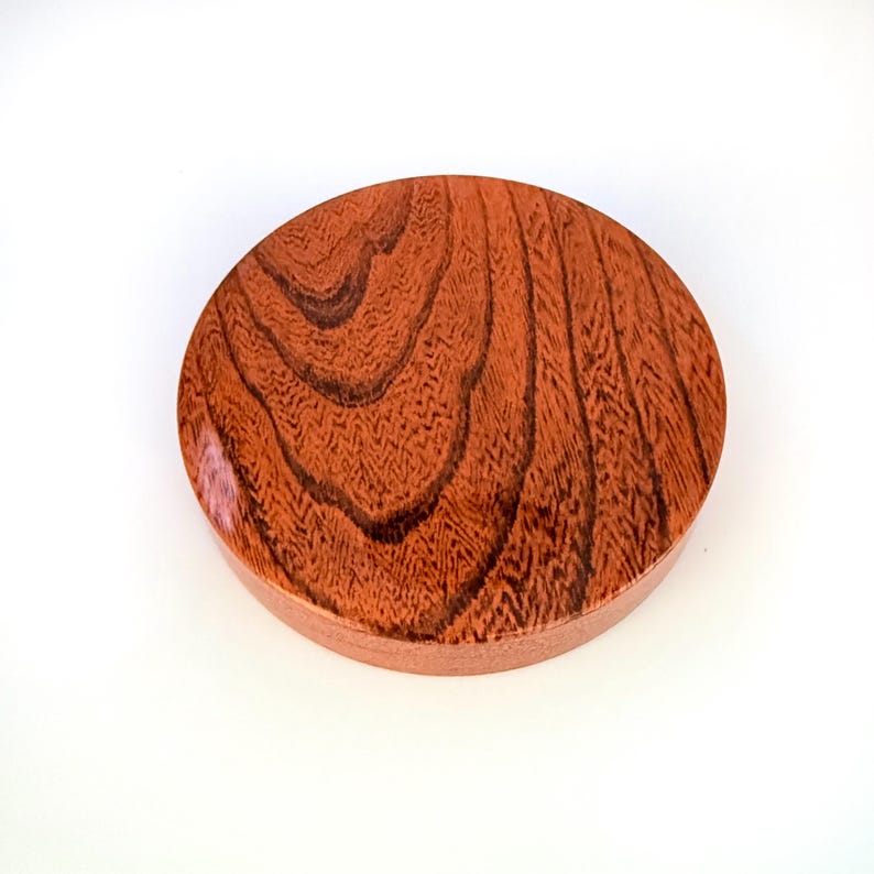 May include: A circular wooden lid with a smooth, polished finish. The surface displays a prominent, swirling grain pattern in shades of reddish-brown and dark brown, creating a natural, organic texture across the entire top of the round object.