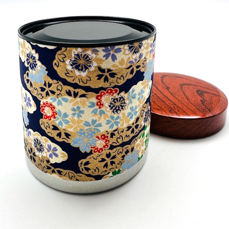 May include: A cylindrical tea canister featuring a dark blue fabric wrap with a gold, red, and light blue floral pattern. The container has a black rim and a separate, circular, polished reddish-brown wooden lid with a visible wood grain texture.