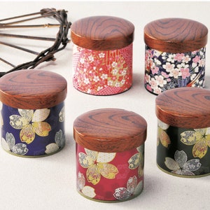 May include: Five cylindrical canisters with dark brown wood-grain lids. Each canister features a distinct floral pattern in red, blue, black, and pink with gold accents. A dark, curved, twig-like decorative element is positioned to the upper left.