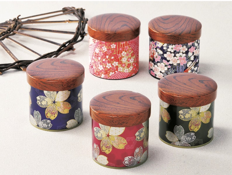 May include: Five cylindrical canisters with dark brown wood-grain lids. Each canister features a distinct floral pattern in red, blue, black, and pink with gold accents. A dark, curved, twig-like decorative element is positioned to the upper left.