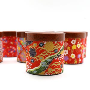 May include: A cylindrical storage canister with a dark brown wood-grain lid and a vibrant red body featuring a detailed pattern of colourful flowers, green foliage, and gold-toned line work. The container has a flat base and a matching circular lid.
