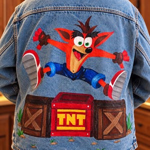 Puede incluir: Una chaqueta de mezclilla azul con una ilustración pintada a mano en la espalda. El diseño muestra al personaje naranja Crash Bandicoot saltando sobre tres cajas de madera, incluyendo una caja roja central con el texto amarillo TNT impreso.