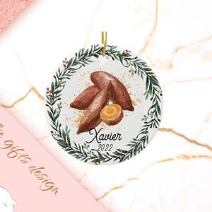 May include: A white ceramic ornament with a green wreath design and a brown sweet potato illustration. The ornament has the name "Xavier" and the year "2022" written on it.