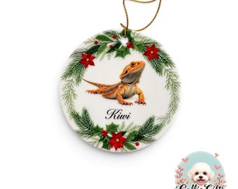May include: A round white ceramic ornament featuring a central image of an orange bearded dragon lizard. The lizard is surrounded by a festive wreath of green pine needles, red holly berries, and red poinsettia flowers. The name Kiwi is printed below.