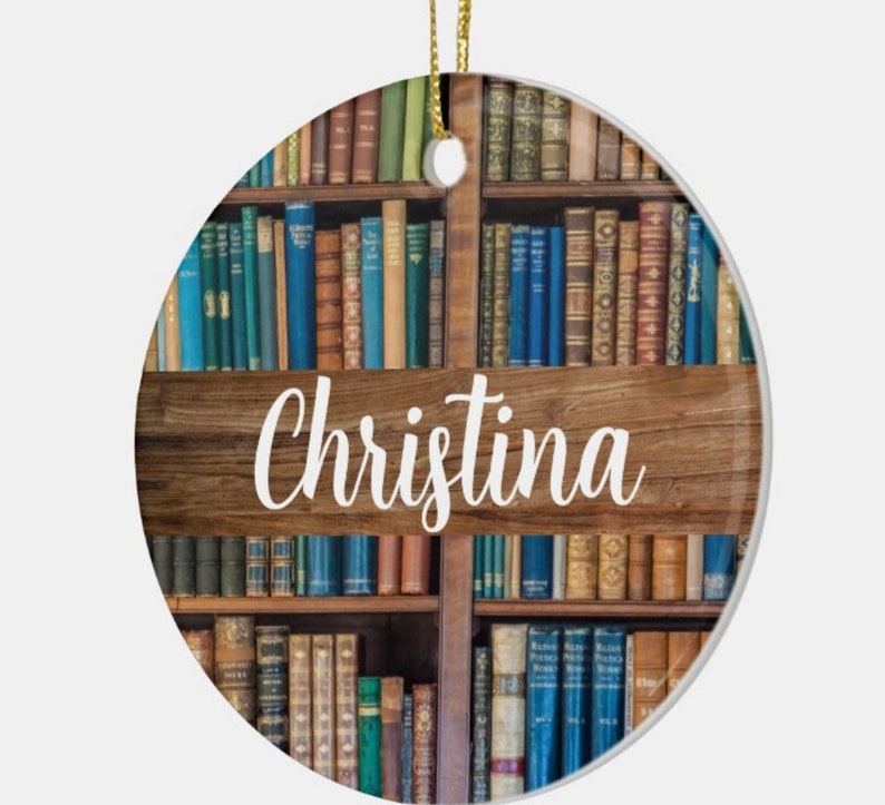 May include: Round ceramic ornament featuring a bookshelf design with various colored books. A wooden sign with the name "Christina" in white script is centered on the ornament. A gold string is attached for hanging.