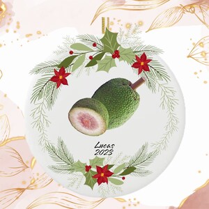 May include: A round Christmas ornament featuring a green breadfruit, one half sliced to reveal the pink interior. The ornament is decorated with a wreath of green leaves, red poinsettias, and the text "Lucas 2023".