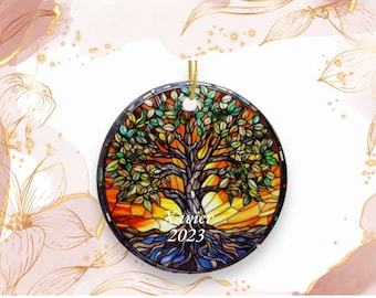 May include: Round Christmas ornament featuring a stained-glass design of a tree with green leaves, a yellow and orange sunset, and blue roots. The name "Xavier" and the year "2023" are printed on the ornament. A gold string is attached for hanging.