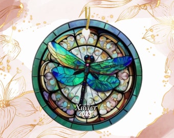 May include: Round ceramic ornament featuring a stained-glass style dragonfly design. The dragonfly has teal and blue wings, a black body, and is set against a background of beige and gold. The ornament has a green border and a gold string for hanging. Text reads "Xavier 2023".