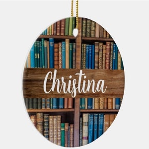 May include: Oval-shaped ornament featuring a bookshelf design with a wooden plank displaying the name "Christina" in white script. The bookshelf is filled with various antique books in shades of blue, green, and brown, with a gold string for hanging.