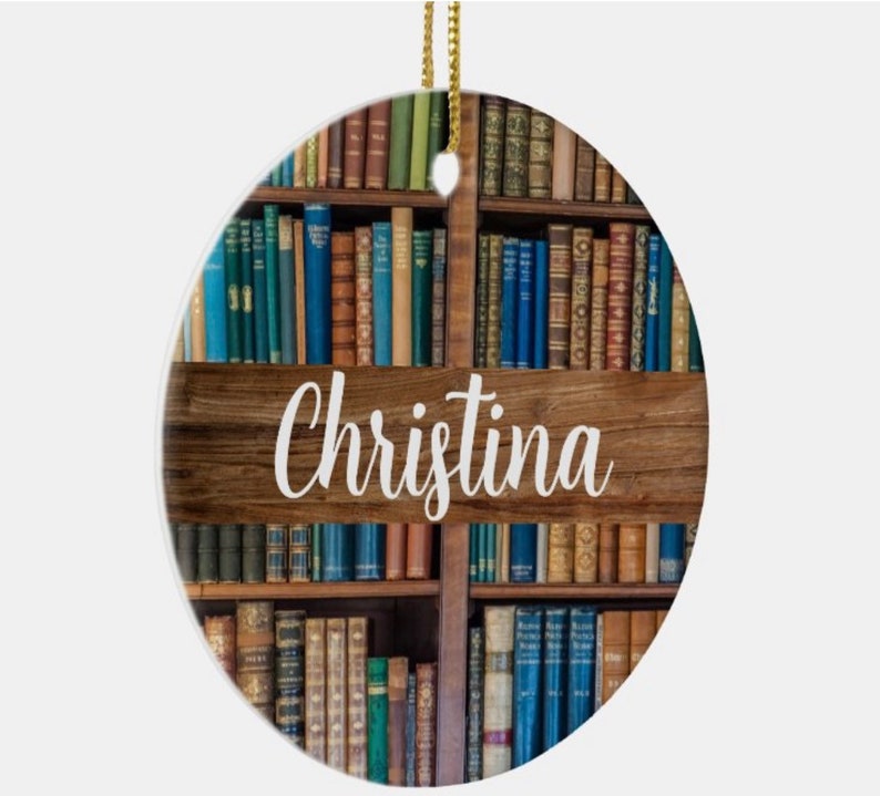 May include: Oval-shaped ornament featuring a bookshelf design with a wooden plank displaying the name "Christina" in white script. The bookshelf is filled with various antique books in shades of blue, green, and brown, with a gold string for hanging.