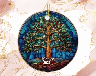 May include: Round ceramic ornament featuring a stained-glass design of a tree with red fruit, green leaves, and a brown trunk. The ornament has a blue background and the name "Xavier" and the year "2023" are printed on the bottom. A gold string is attached for hanging.
