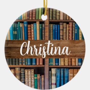May include: Round ceramic ornament featuring a bookshelf design with various colored books. The name "Christina" is written in white script across a wooden beam. A gold string is attached for hanging.