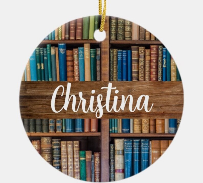 May include: Round ceramic ornament featuring a bookshelf design with various colored books. The name "Christina" is written in white script across a wooden beam. A gold string is attached for hanging.