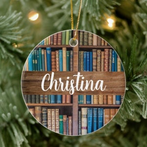 May include: Round Christmas ornament featuring a bookshelf design with various colored books. The name "Christina" is written in white script across a wooden plank. A gold string is attached for hanging.
